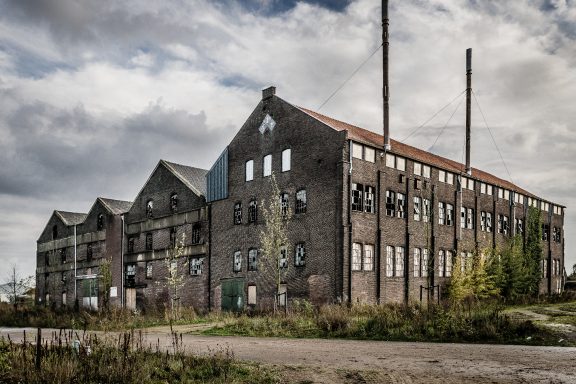 Verlassene Fabrik mit Ziegelmauer, hohen Fenstern und zwei Schornsteinen unter bewölktem Himmel.