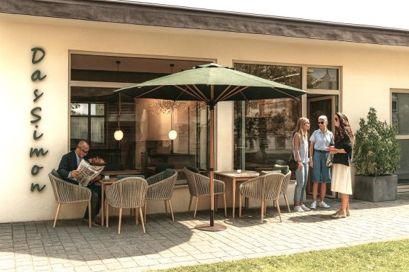 Personen stehen vor einem Café mit Sonnenschirm und Sitzgelegenheiten im Freien.